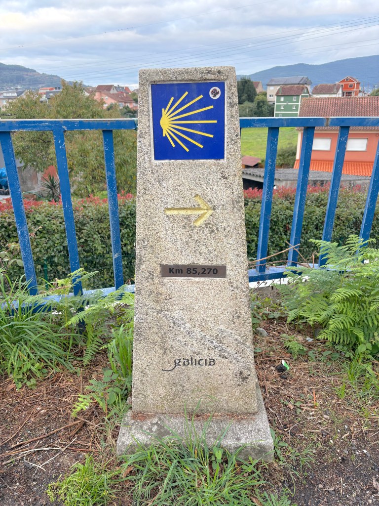 A Camino de Santiago (Portuguese) stone marker indicating 'Km 85,270' with a yellow sunburst symbol, located in Galicia, surrounded by greenery and residential buildings in the background.